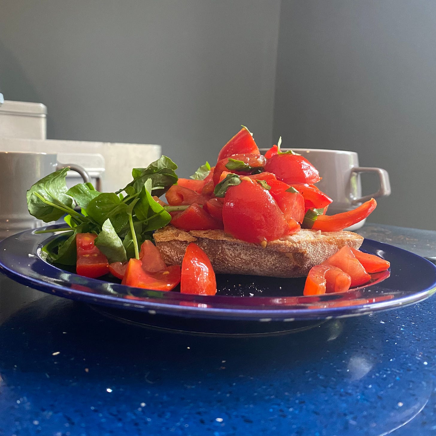 blue plate piled with ciabatta toast, fresh tomatoes, basil and salad leaves. Plate is placed on a blue worktop with a coffee cup behind.