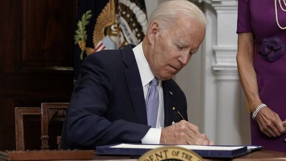 US President Joe Biden signs into law a gun control bill at the White House in Washington DC. He is sitting down, wearing a navy blue suit and tie, with a pen in hand signing the bill. A flag is behind him and a woman in a purple dress and pearls stands beside him.