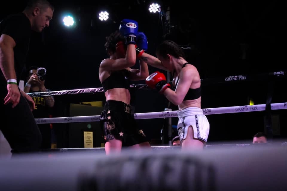 Female fighter in white shorts and black sports bra punches a female fighter in black shorts and sports bra, ref watches closely