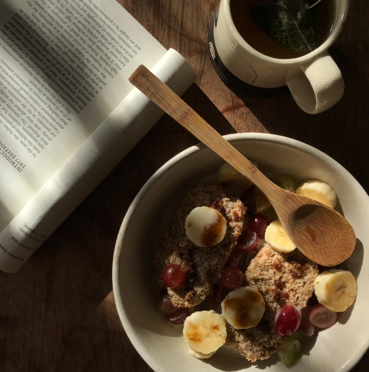 A photo in bright morning light, taken from above. A bowl of malty oat bars in the bottom right, with sliced rounds of banana, red grapes, and a bamboo wood spoon placed over. Above a full mug of mint tea. To the top left, cut off by the frame, is book rolled open to a central page, the text upside down and illegible. 