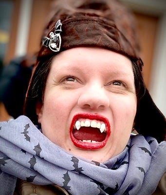 Portrait of Jennifer Lee Rossman. She is wearing a brown hat with a decal in the shape of a silver winged potion bottle, and a purple scarf with a pattern of bats. She is baring her teeth in an exaggerated snarl, showing off a pair of costume vampire fangs.