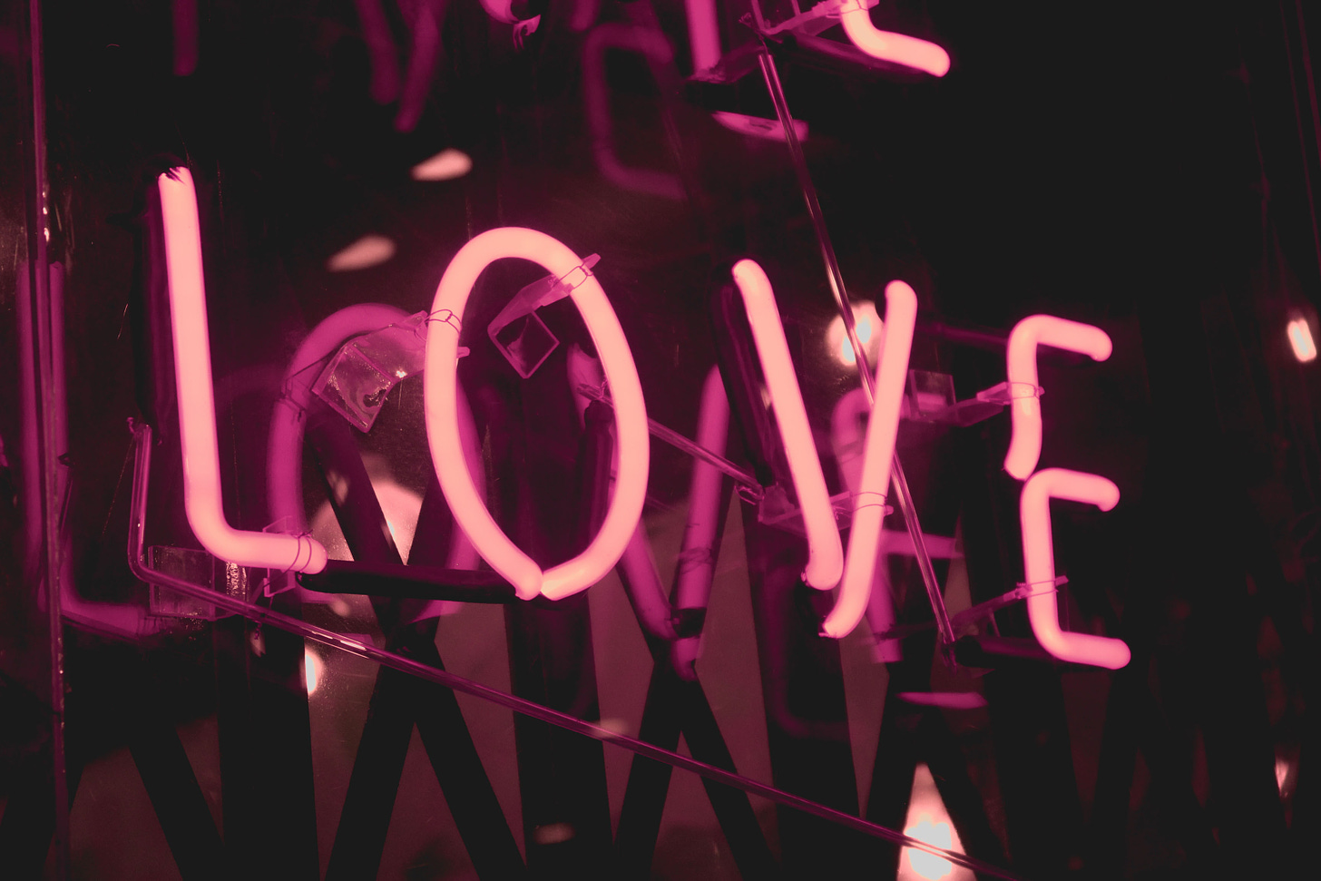 Titelbild: Nahaufname eines Neon-Schriftzugs des Wortes "Love" in Rosa und Lila mit dunklem Hintergrund. Zu sehen ist auch seine Aufhängung an einer Hauswand, in einem Club oder dergleichen. Bild: Shaira Dela Peña via Unsplash