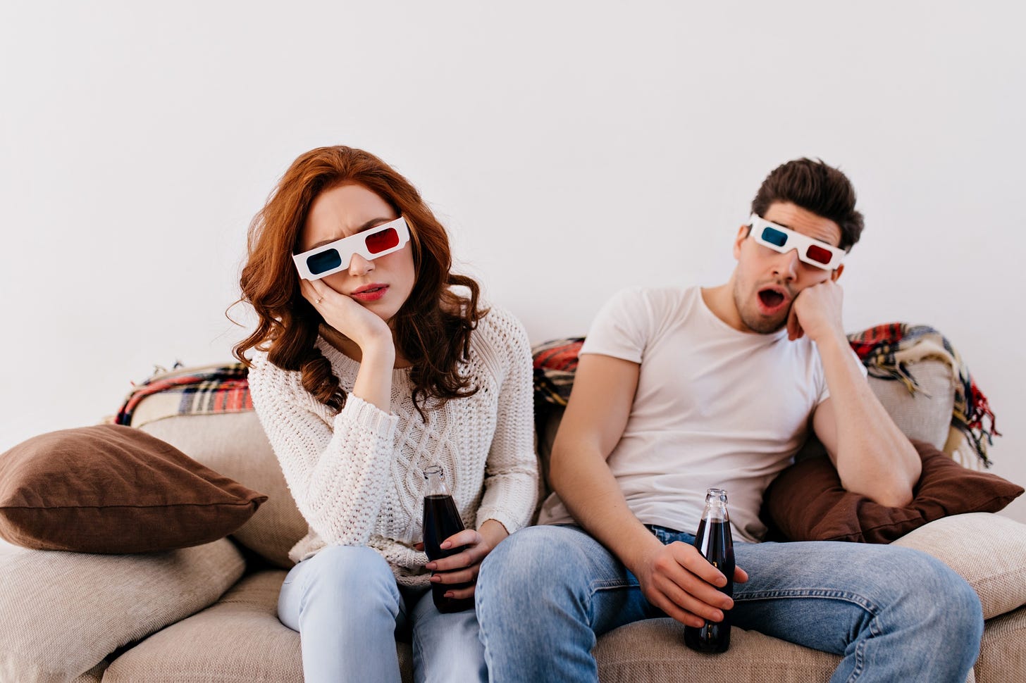 A bored couple sits on the couch drinking soda and wearing 3D glasses.