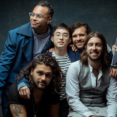 A photo of the band, five men wearing a range of casual and semi-casual clothing, huddled together at varying heights against a dark background and smiling at the camera. 