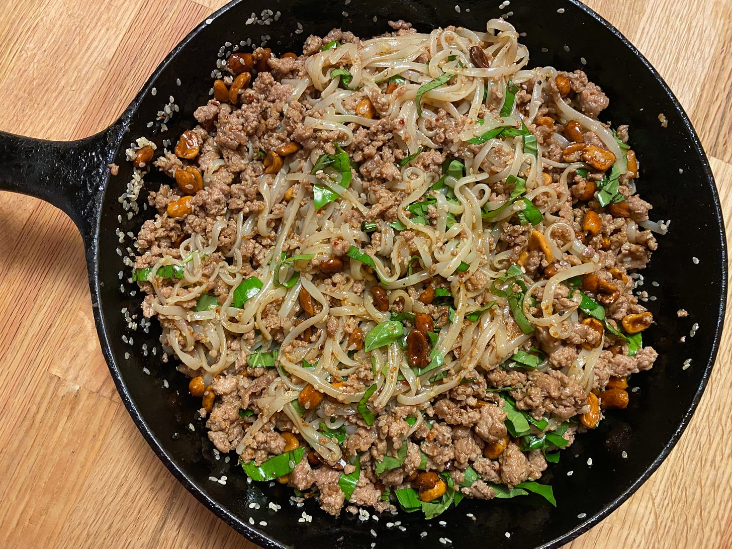 A cast iron frying pan of rice noodles scattered with ground pork, peanuts, and chopped basil.