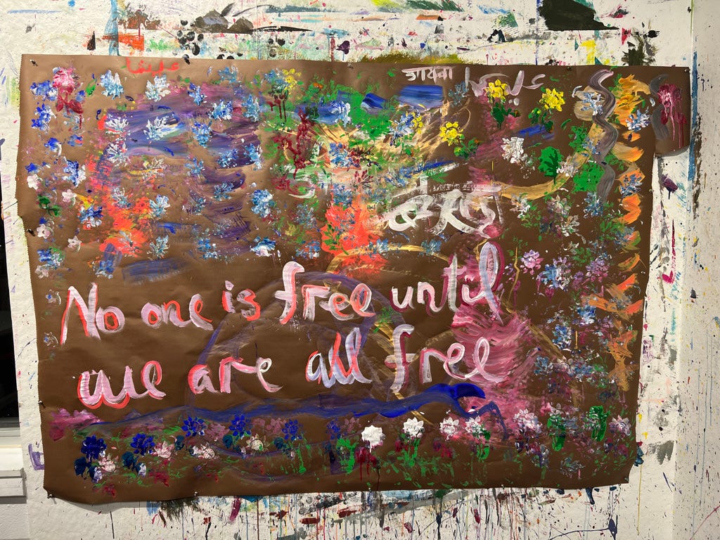 A spontaneous, unplanned, vibrant, multi-colored, chaotic, free-style painting Ayesha made while blind-folded using multiple tools- from paint brushes to stencils to hands, with the words “No one is free until we are all free” prominently written on it in English & much smaller size in Urdu, Arabic, Hindi and Kannada (mostly covered by paint).