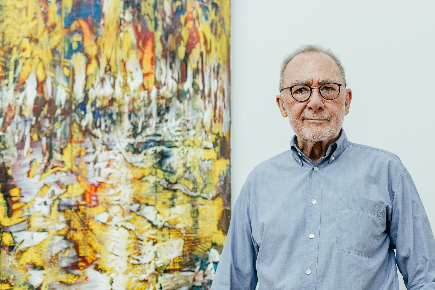 A portrait of Gerhard Richter in a blue shirt standing next to one of his paintings