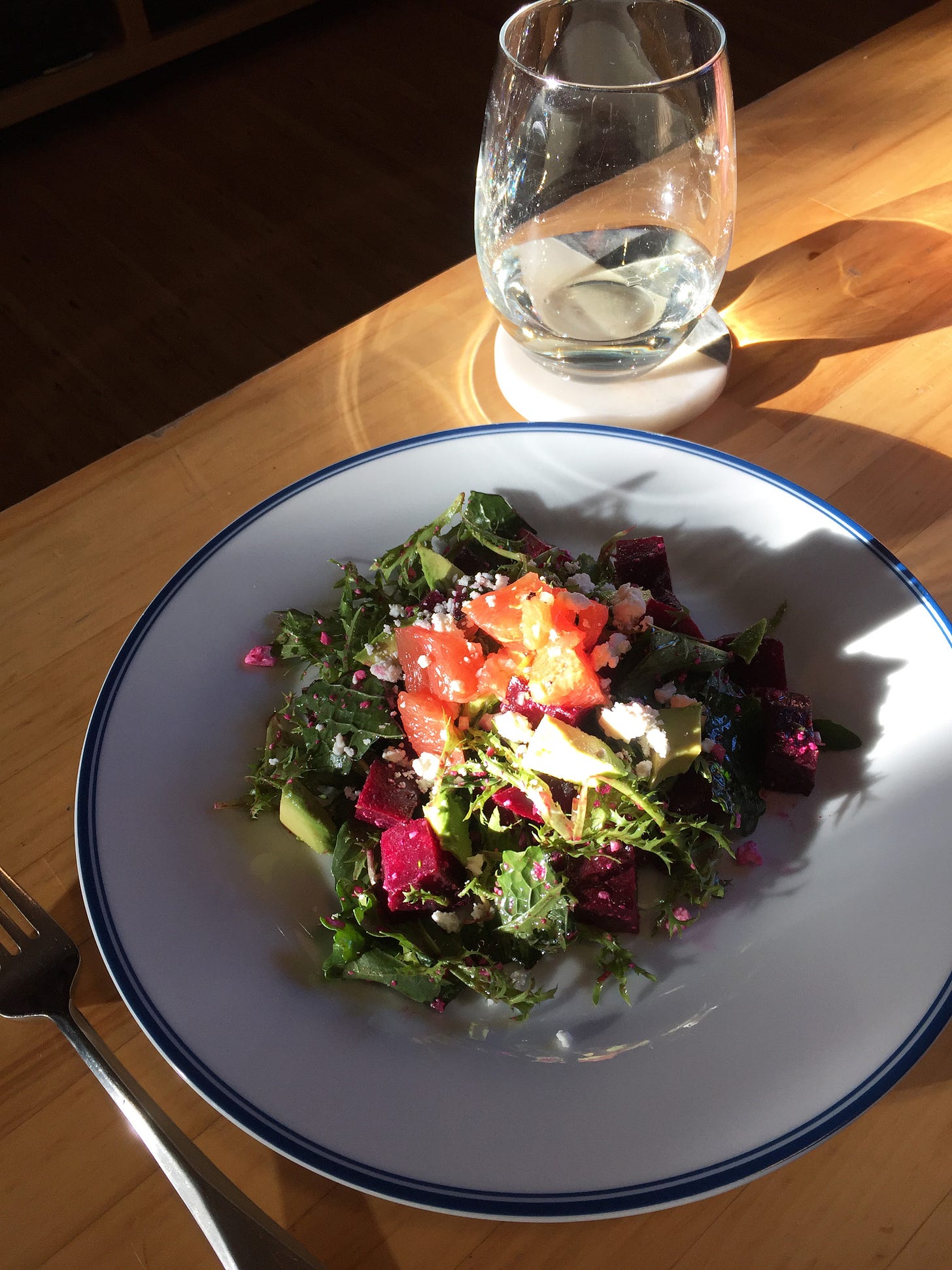 a plate of beet salad with dark greens, avocado, crumbles of feta, and grapefruit segments on top. The plate site in a patch of sun on a wooden table with a glass of white wine at the upper corner.