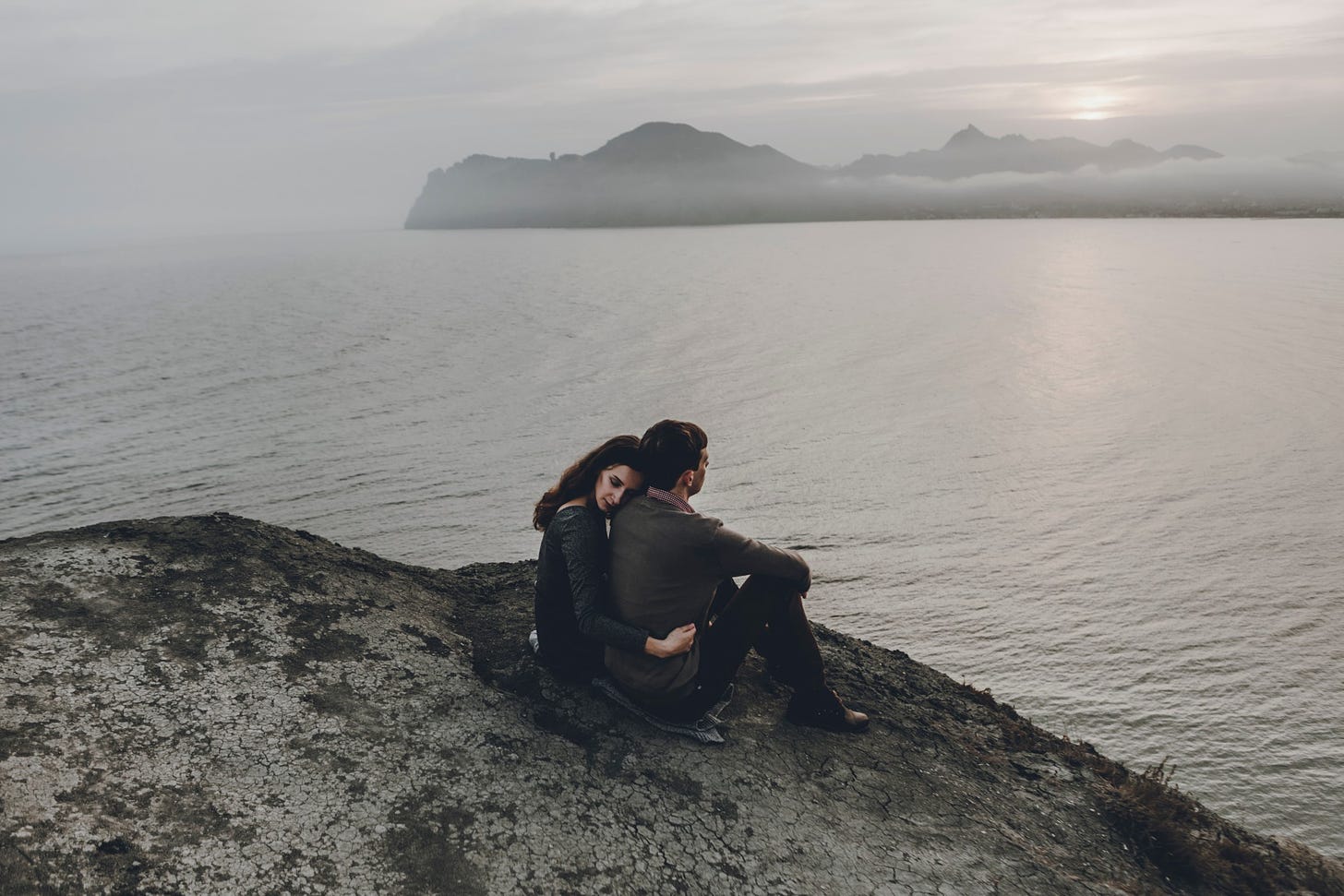 A couple embraces on a cliff overlooking a grey and cloudy sea.