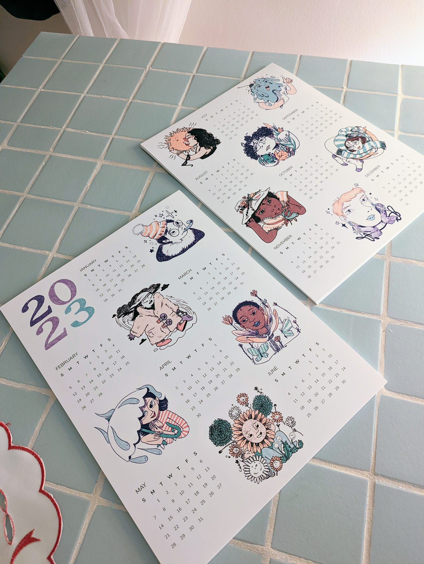A photo of the front and back of the Litte BBs calendar for 2023. Strange and cute seasonal characters accompany each month of the year. The calendars sit on top of a light blue tiled dining table.