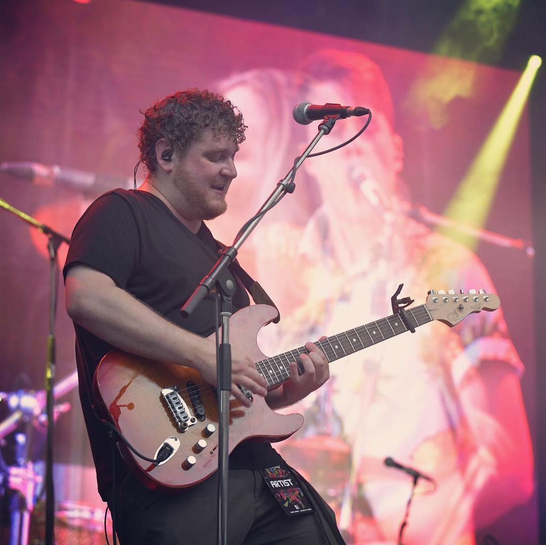 Eric Hitsman playing the guitar on a stage in front of a screen with a projection of his bandmates.