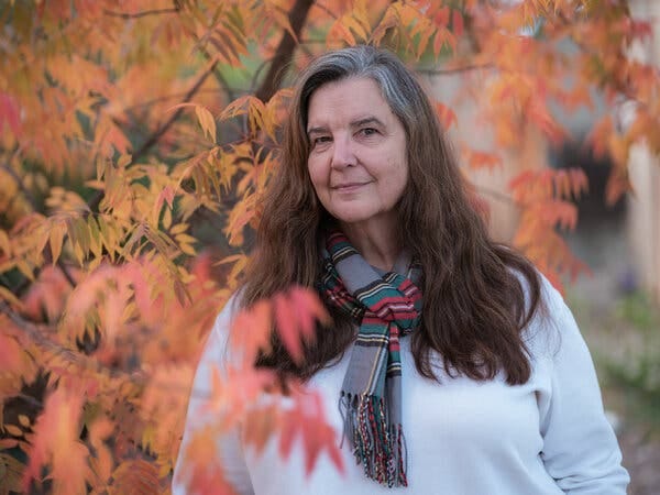 A white woman with long brown-and-gray hair wears a white shirt with a plaid scarf around her neck. She is standing in front of a tree with leaves that have started changing color for the fall.