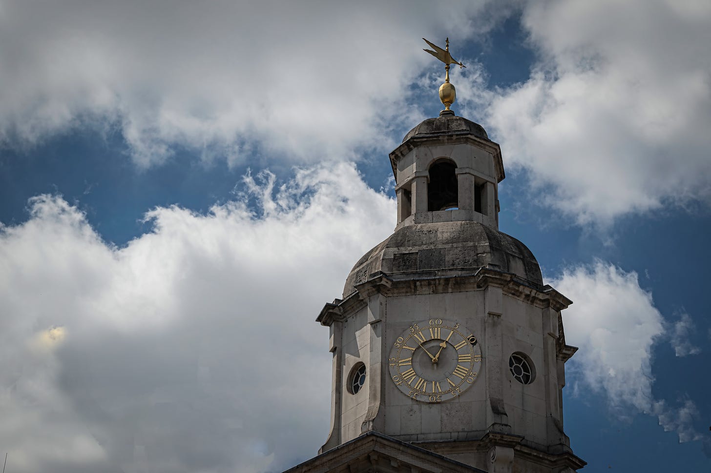 The tower of a building in London with a clock with  Roman numeral sat on one side, the time 12:52 and a weather vane on top  against a blue sky and large fluffy white and grey clouds