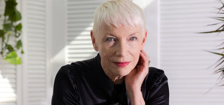 A woman with short white hair half smiling with one hand cupping her face