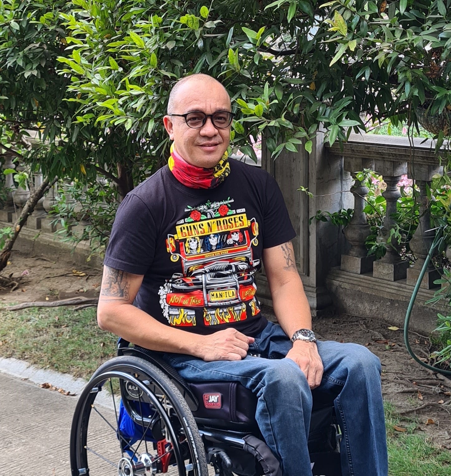 A photograph of Abner, a Phllippinian man, sitting in a wheelchair, half-smiling in a knowing way towards camera. Abner has dark circular glasses and is wearing jeans with a black Guns N’ Roses Manila t-shirt and a neck-scarf in red and yellow colours matching the t-shirt illustration. He has tattoos on his upper arms, and the background is green vegetation with an elegant stone wall. 