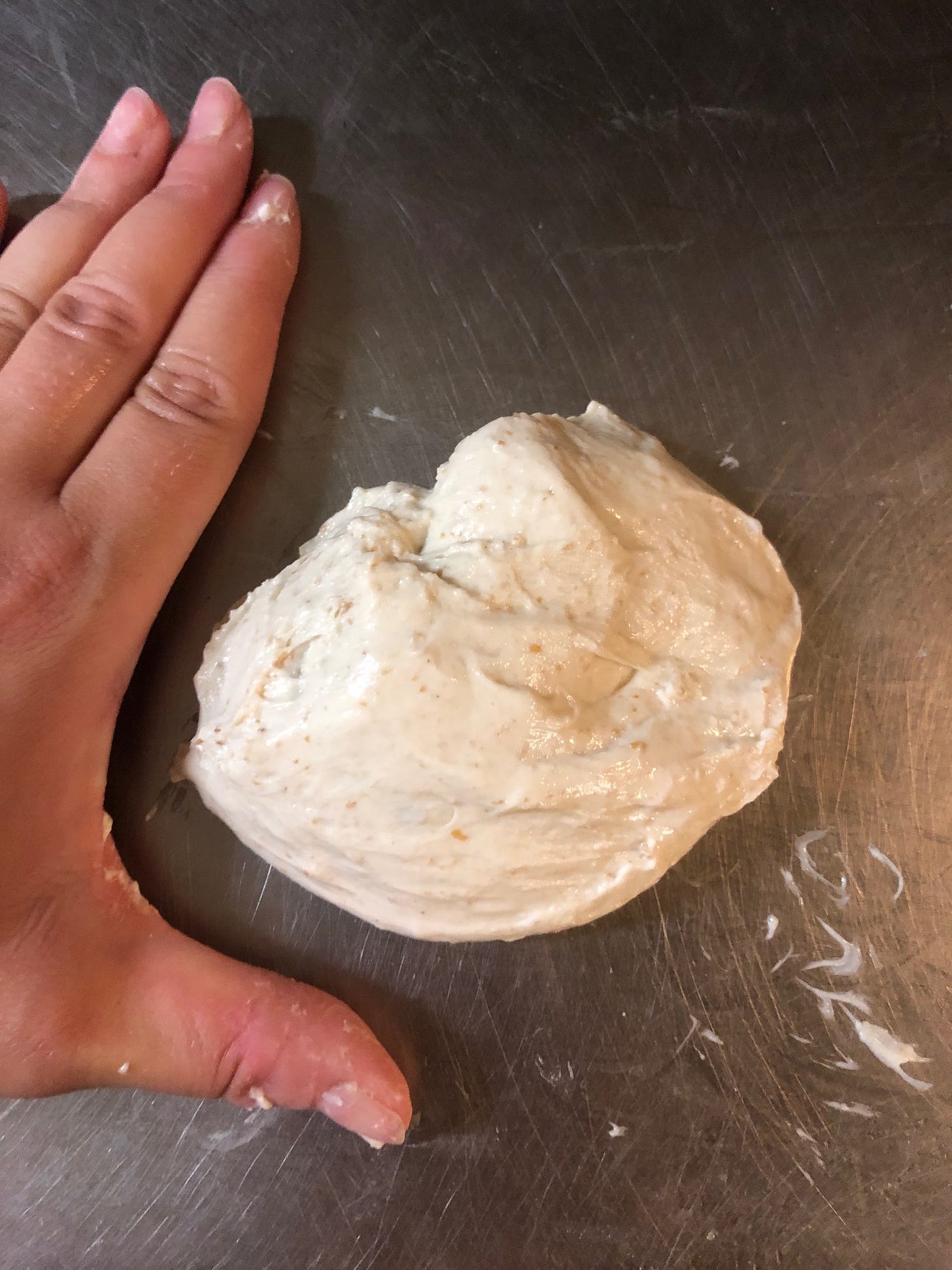 Left hand, palm down, on a metal table next to a piece of raw unshaped dough.