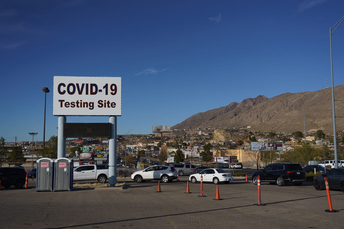 Under a blue sky and a sign advertising Covid-19 testing, cars line up. There are mountains in the background.