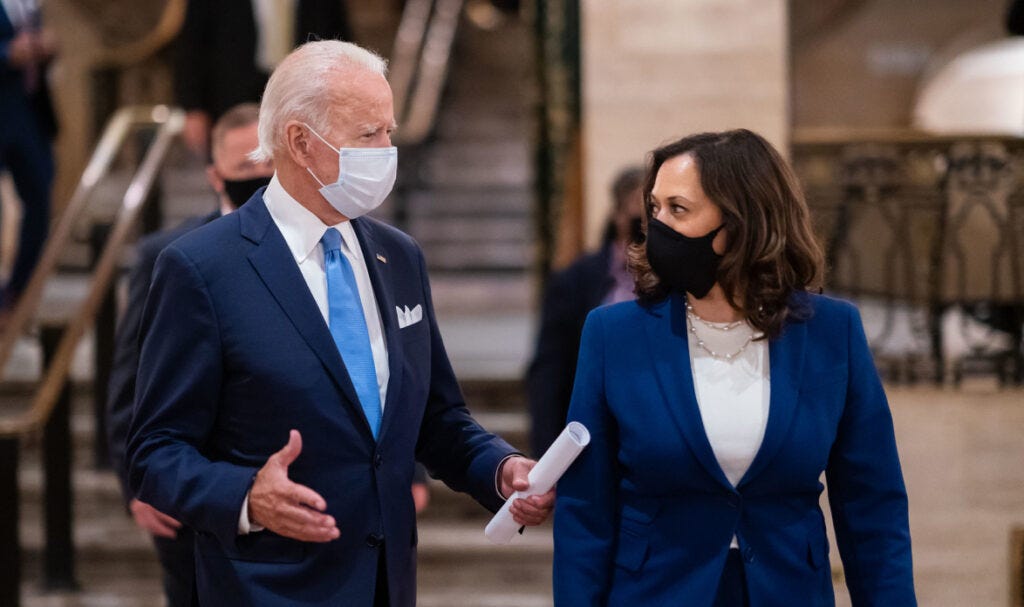 Joe Biden and Kamala Harris walking through the lobby of a building. They are both wearing suits and masks. Joe is holding some rolled up papers in one hand.