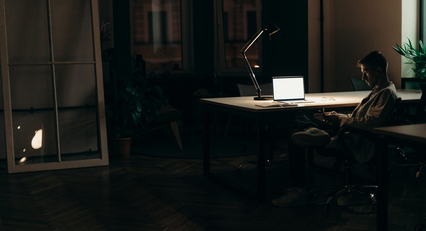 man sitting at desk in dark room with lamp on on the desk