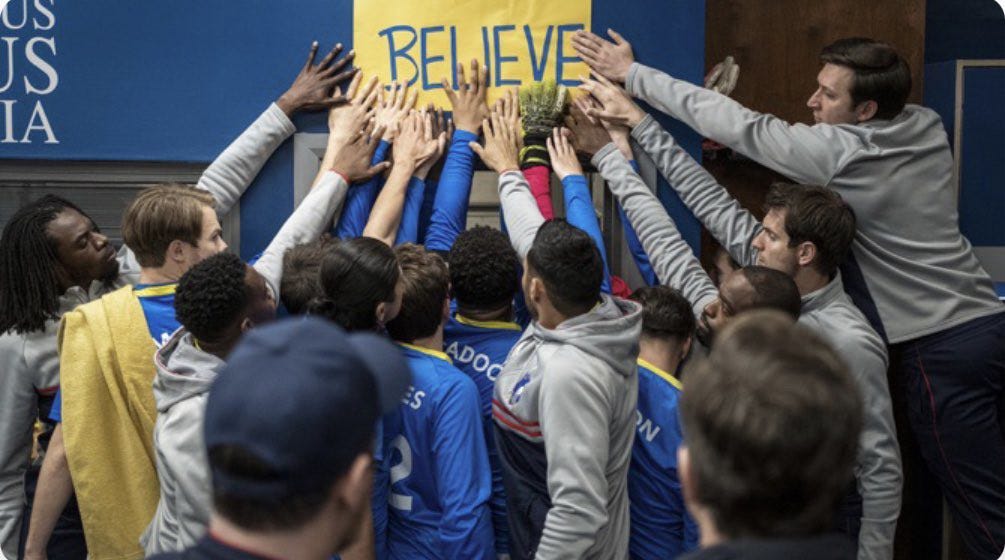 Soccer team from Ted Lasso touching a yellow sign that says BELIEVE