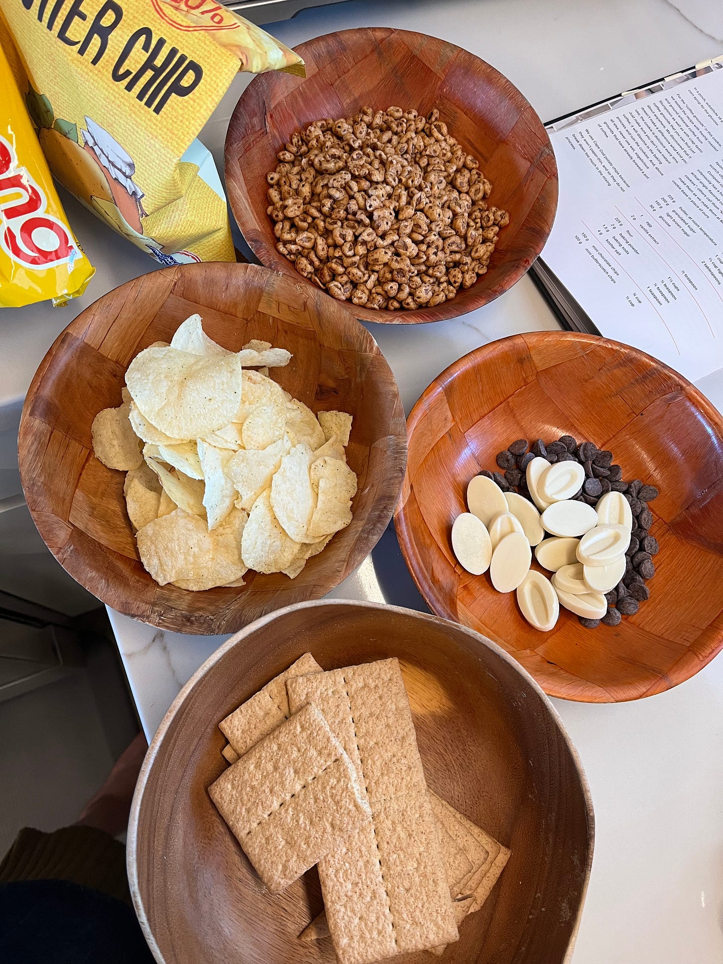A kitchen counter with bowls filled with snacks: Jolly Pong puffed grain snacks, potato chips, chocolate chips, and graham crackers.