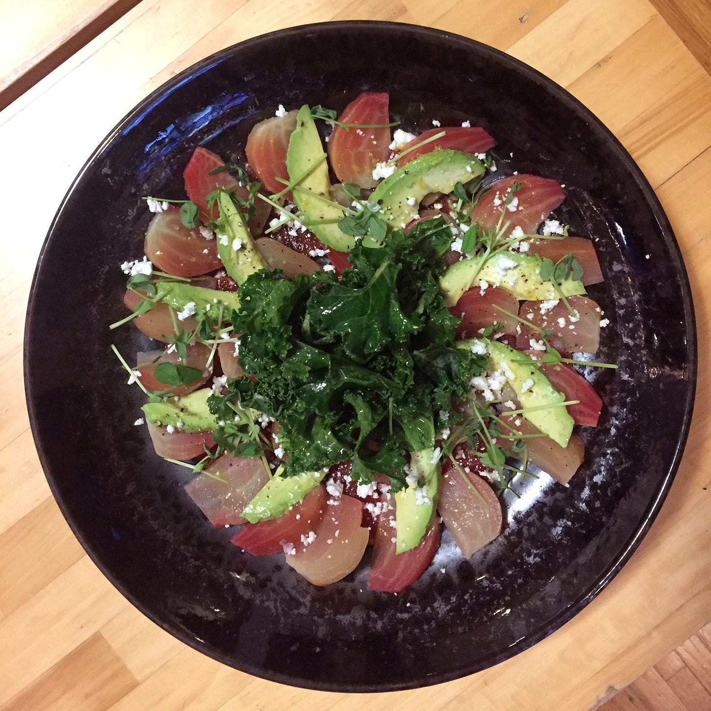 a shallow black bowl filled with slices of pink beets arranged with avocado in a circle. Slices of blood orange are just visible below them, and on top is a small pile of kale leaves and a sprinkling of feta and pea shoots.