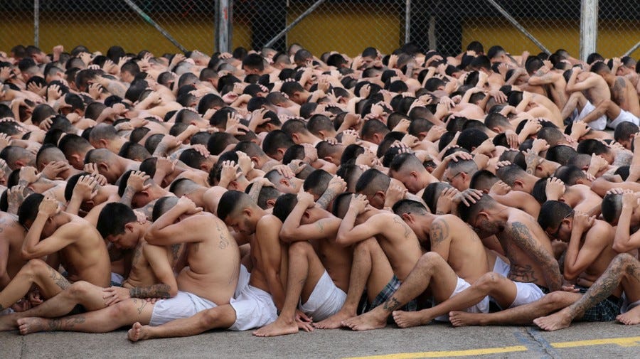 Dozens of men wearing only boxer shorts are gathered in a courtyard, seated on the ground close to one another, with their hands on their heads.