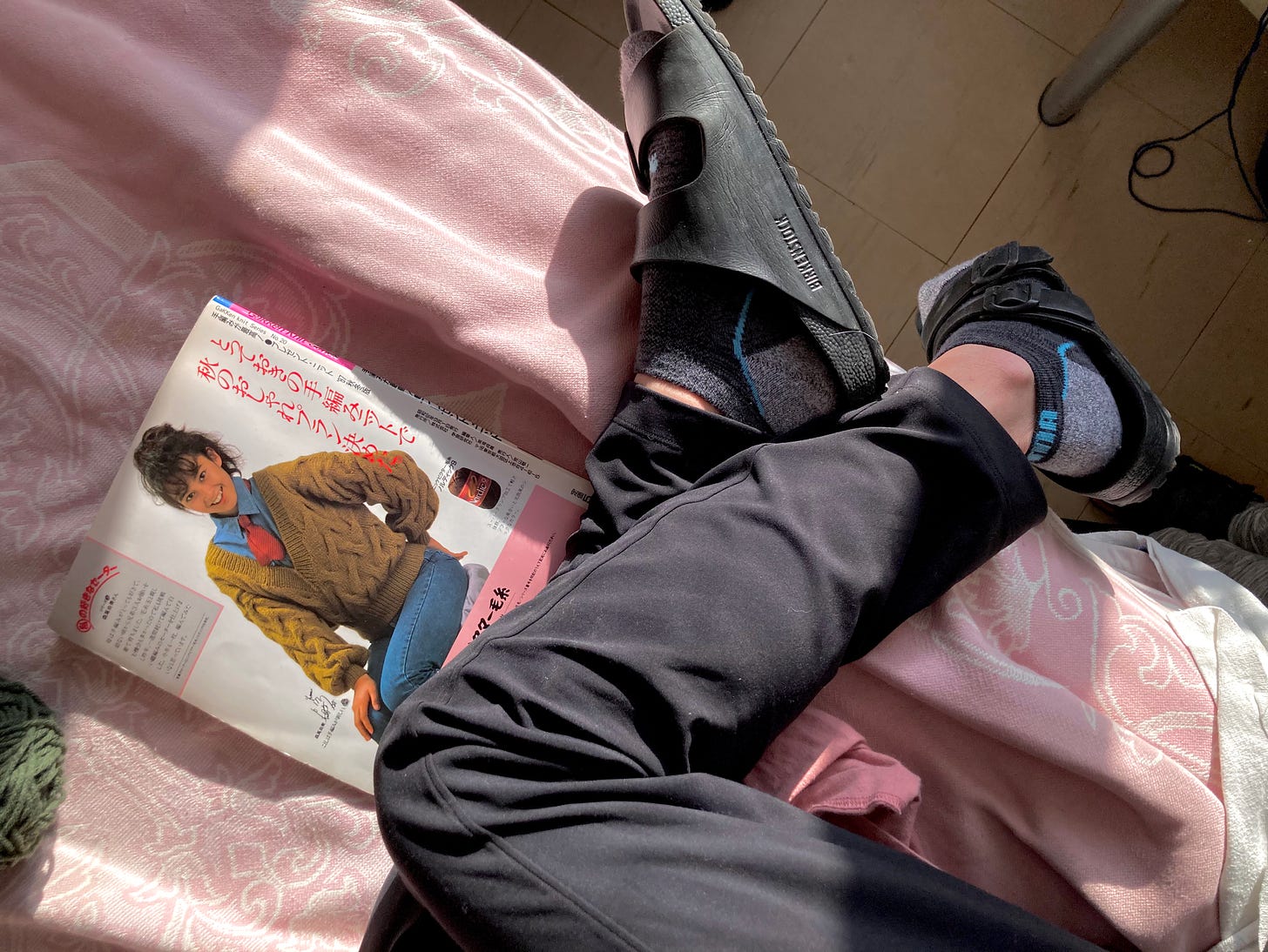 Aerial view of legs in black pants with black birkenstocks on a bed with a pink blanket. 