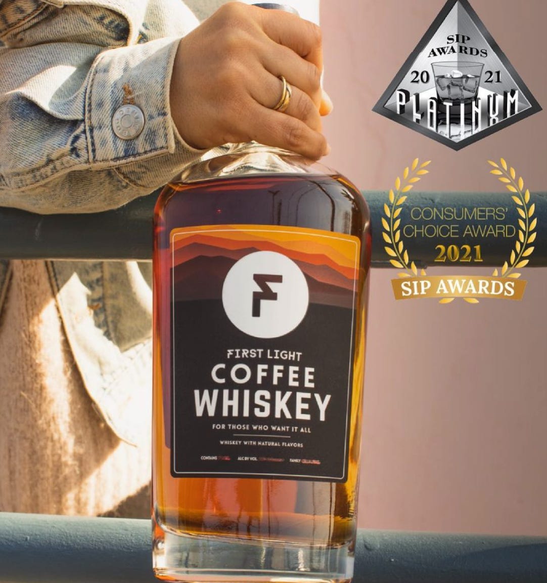 A bottle of First Light Coffee Whiskey held up close to the camera by a man's hand with a gold ring on the ring finger, and a stone-washed jean jacket visible to just past the cuff.