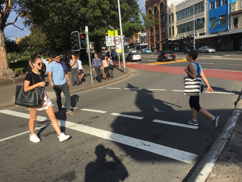 A signalized but porkchop-islanded crosswalk at a Free Left (Free Right for those in the right-side drive countries). Notice the pedestrian light is red (don't walk) but the pedestrians cross anyway. If the free left is not eliminated in a more comprehensive redesign, it could easily be de-signaled and the crosswalk raised, so pedestrians dominate, and cars travel when they can.