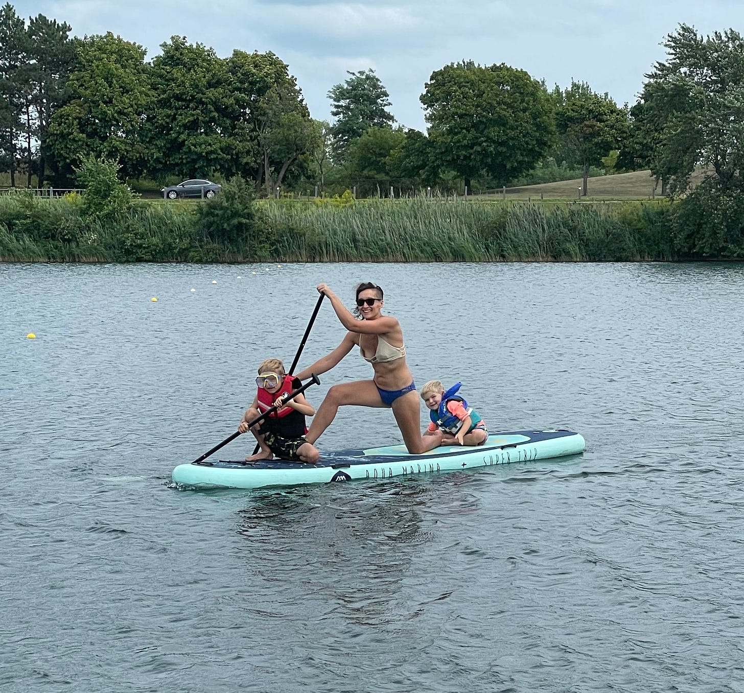 Tarzan and her two young boys on a stand up paddleboard, her seven year old son clumsily trying to paddle and her three year old dipping his fingers into the water at the back. the three of them look really happy