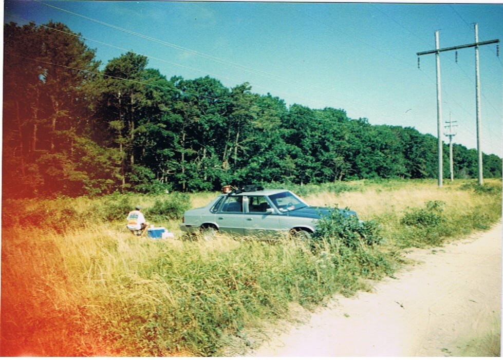 An actual photograph of my 1985 Plymouth Caravelle parked behind some bushes in the power lines on the Cape, below a depthless slate blue sky fading to white at the edges. Behind it my friend Bill squats next to a blue cooler, which probably has melted ice and a couple of rapidly warming steaks in it.