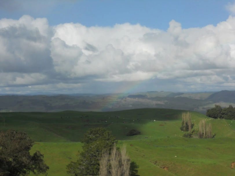 The rolling green hills of the north island of New Zealand. Above is blue sky filled with puffy clouds. In the middle of the photo is the hint of a rainbow.