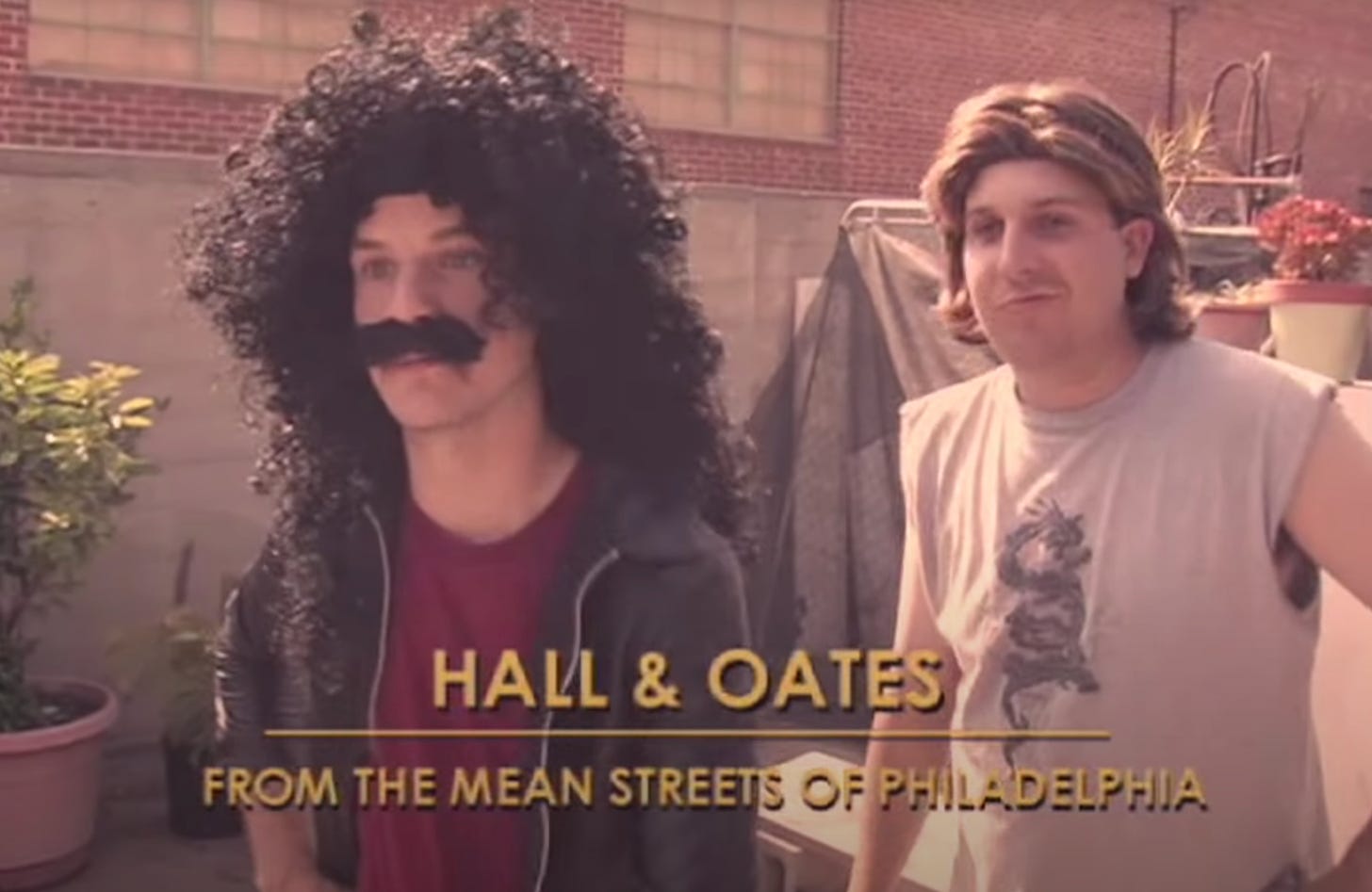 man in a long black curly wig with a fake moustace. Behind him is a man with a blonde wig. The caption reads "Hall and Oates - from the mean streets of Philadelphia"