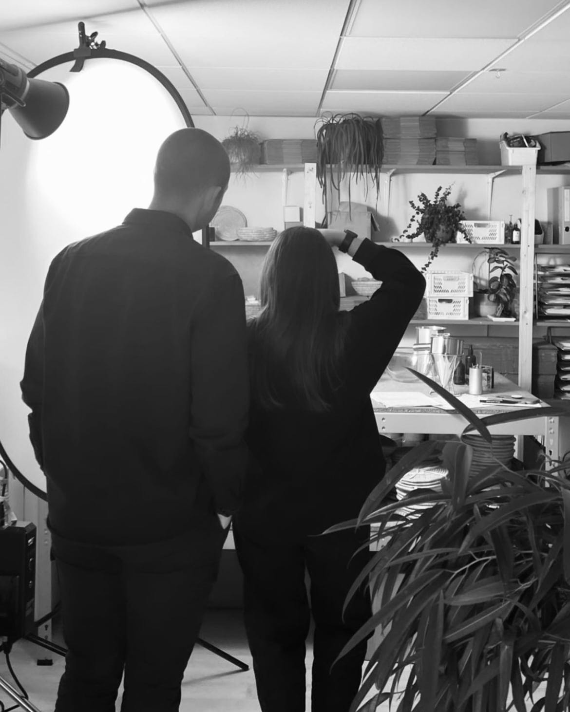 A man and a woman have their back to the camera. They are taking pictures in a workshop with a big light to their left and shelving on the right.