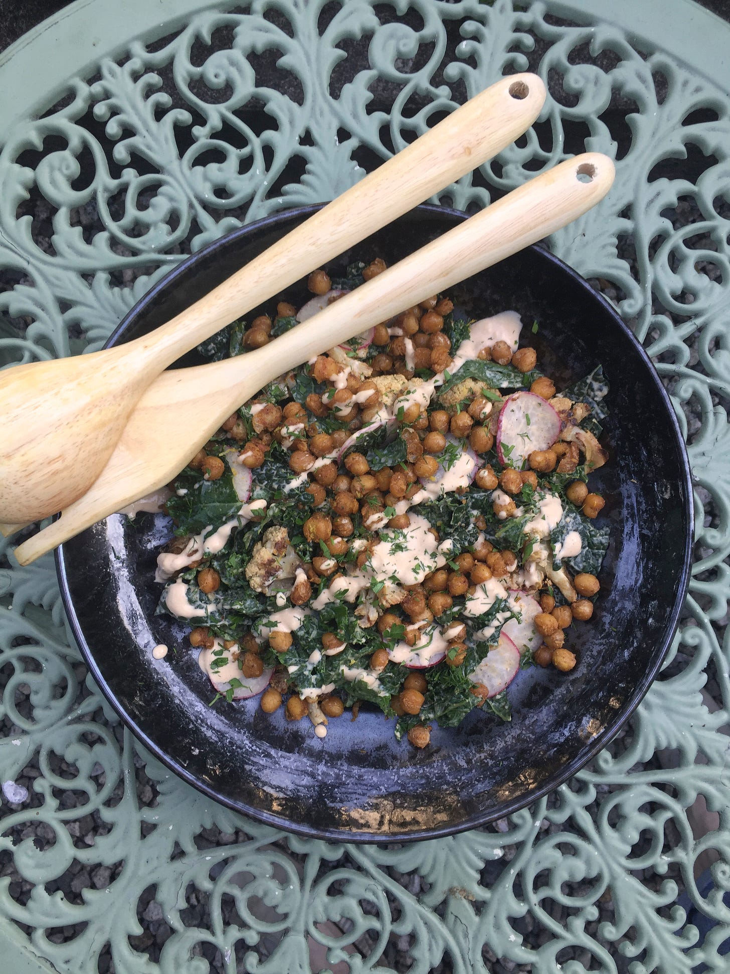 on a wrought-iron outdoor table, a shallow black dish of kale and roasted cauliflower with chickpeas and herbs and a drizzle of tahini dressing overtop. Wooden salad utensils rest across the top edge of the dish.