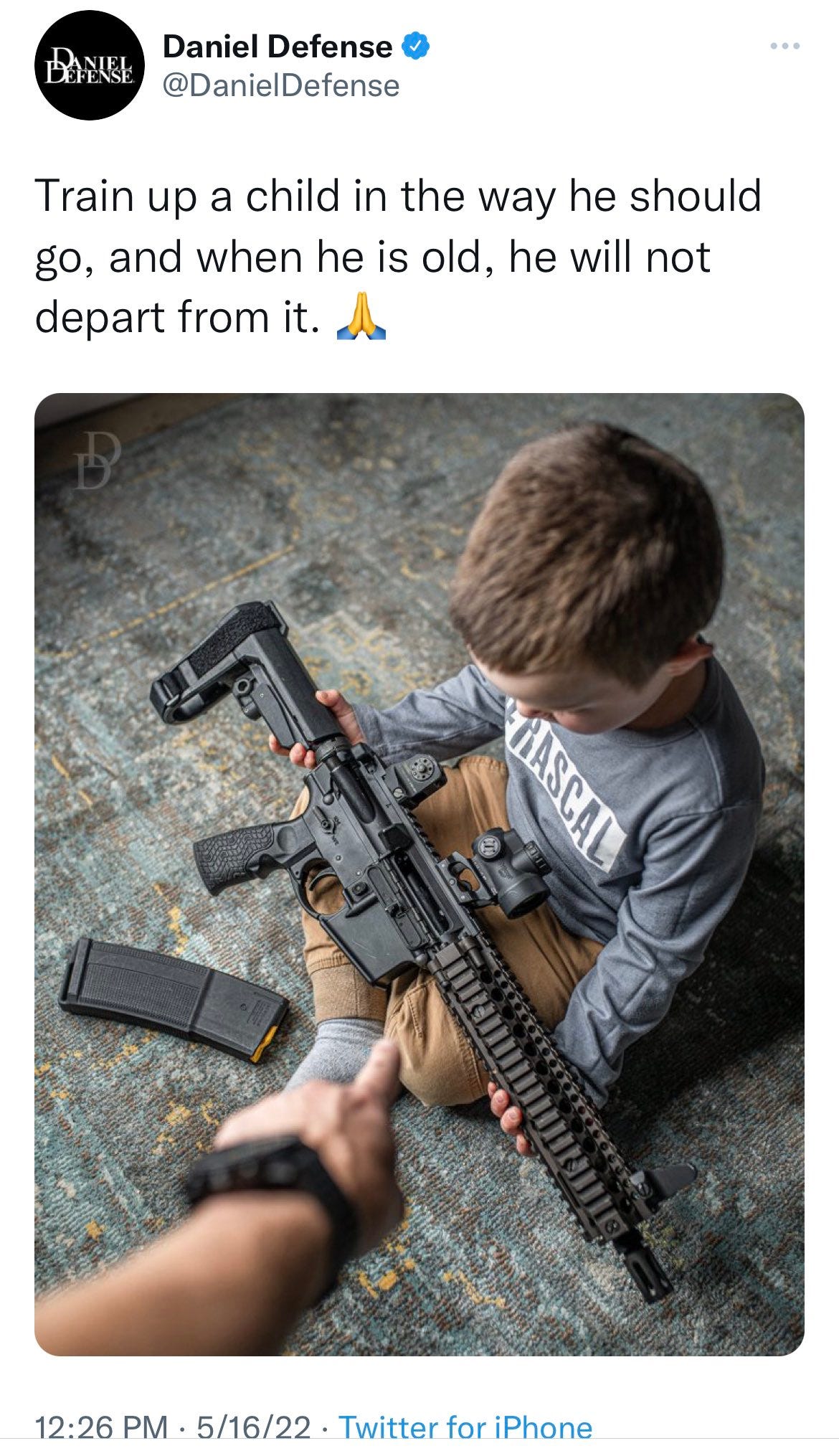 Screenshot of a tweet by @DanielDefense dated 5/16/22 that reads “Train up a child in the way he should go, and when he is old, he will not depart from it. 🙏” with a picture of an adult hand in the close foreground pointing down at a boy, maybe 6 years old, sitting cross-legged with an AR-15 style rifle in his lap and a full magazine of rounds on the floor in front of him. 