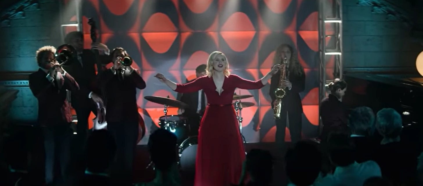 This image shows Agnes Jurati (played by Alison Pill) standing on stage and singing Pat Benatar's 1982 single "Shadows of the Night." Agnes wears a resplendent red dress and spreads her arms wide while belting out the song. This image shows Agnes Jurati (played by Alison Pill) standing on stage and singing Pat Benatar's 1982 single "Shadows of the Night." Agnes wears a resplendent red dress and spreads her arms wide while belting out the song.