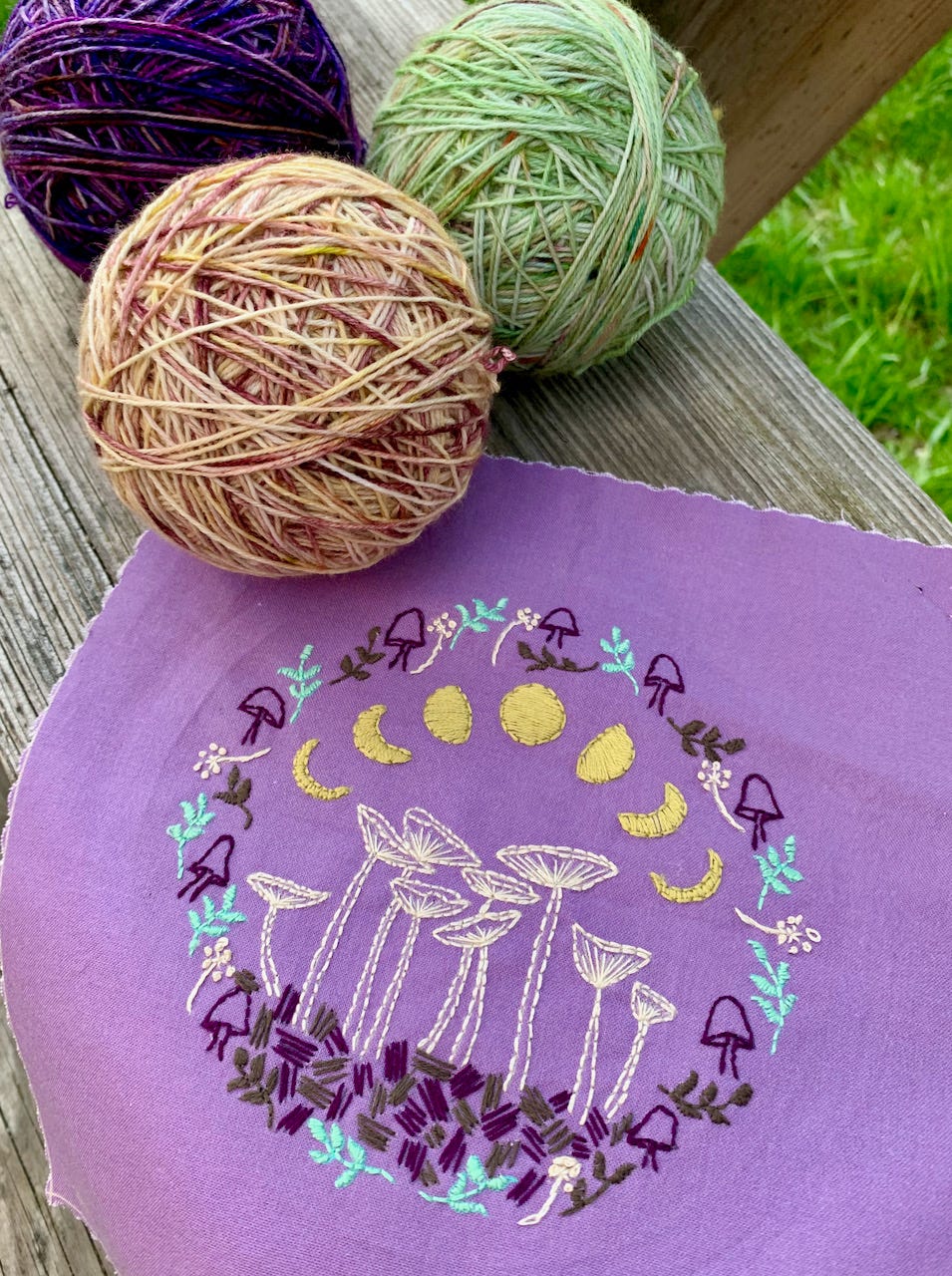 Three balls of yarn purple green peach, and a finished hand embroider piece of moon phases and mushrooms