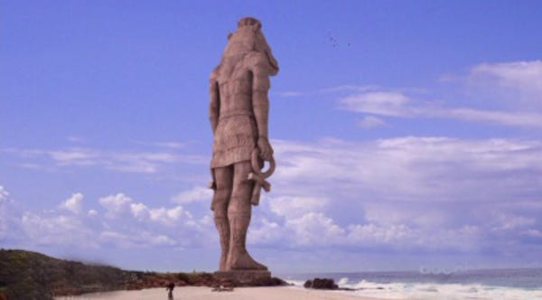An immense statue of the Egyptian god Tawaret stands on a beach, looking out over the water.