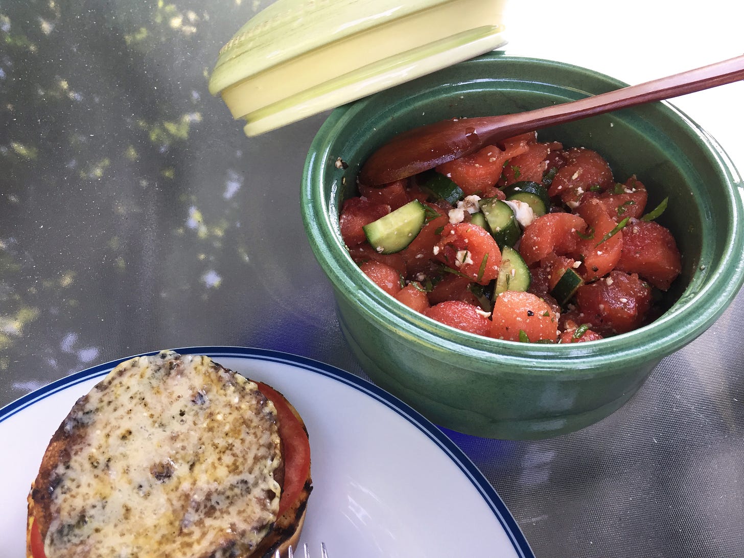 a green bowl of watermelon and cucumber salad with herbs and feta. A wooden spoon sits in the bowl and the bowl's lid rests on its edge. In the bottom corner, an open-faced cheeseburger is just visible.