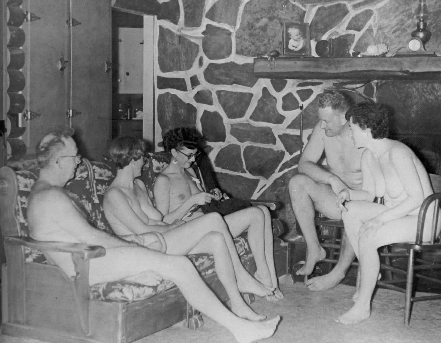 Black and white photo of five adult nudists, men and women, sitting together in a living room. By photographer Norm Cook, c. 1954