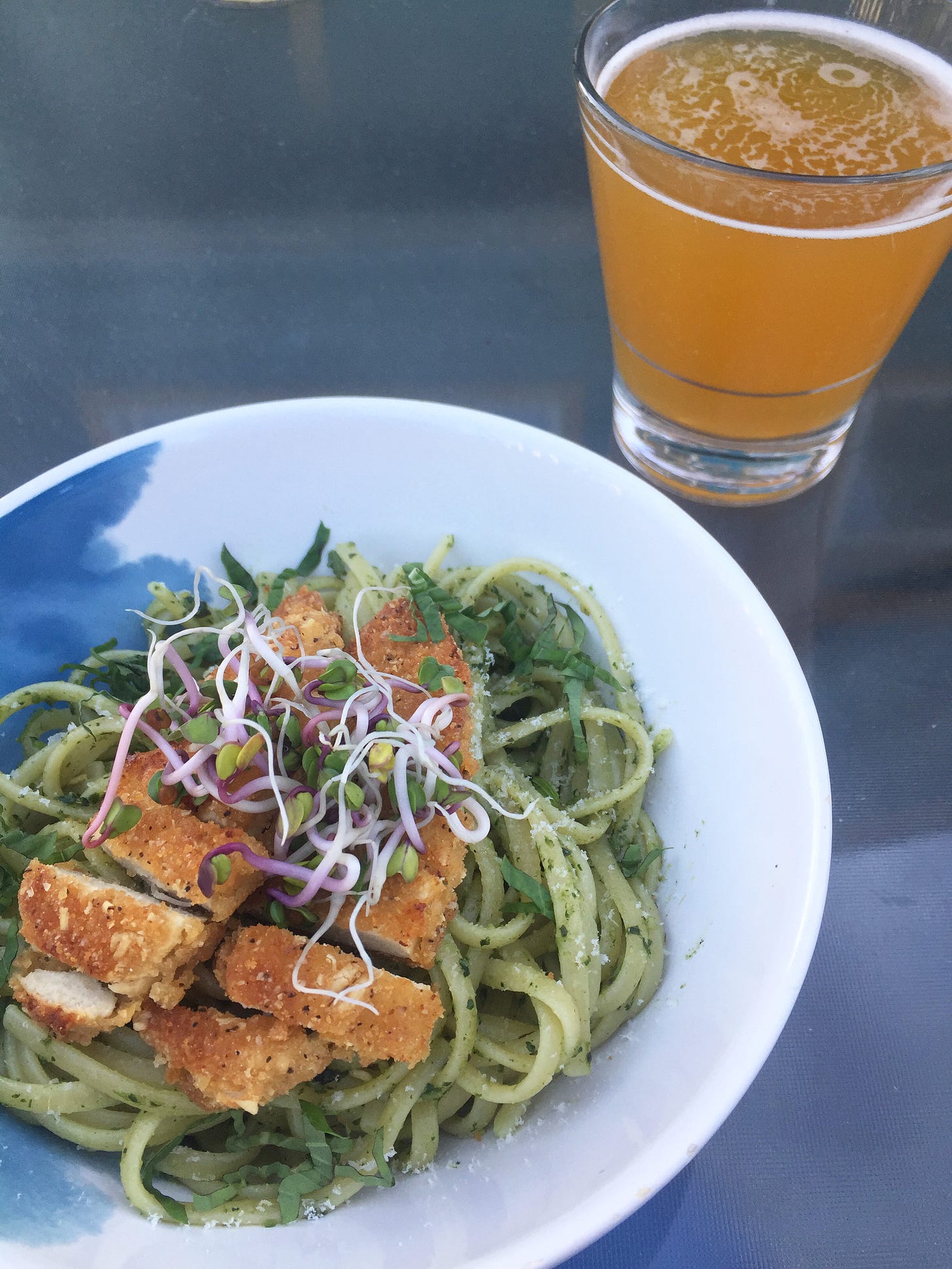 in the foreground, a bowl of linguine in pesto sauce with ribbons of basil and sliced chick'n tenders on top, radish sprouts resting in the centre. a glass of pale beer sits in the background.