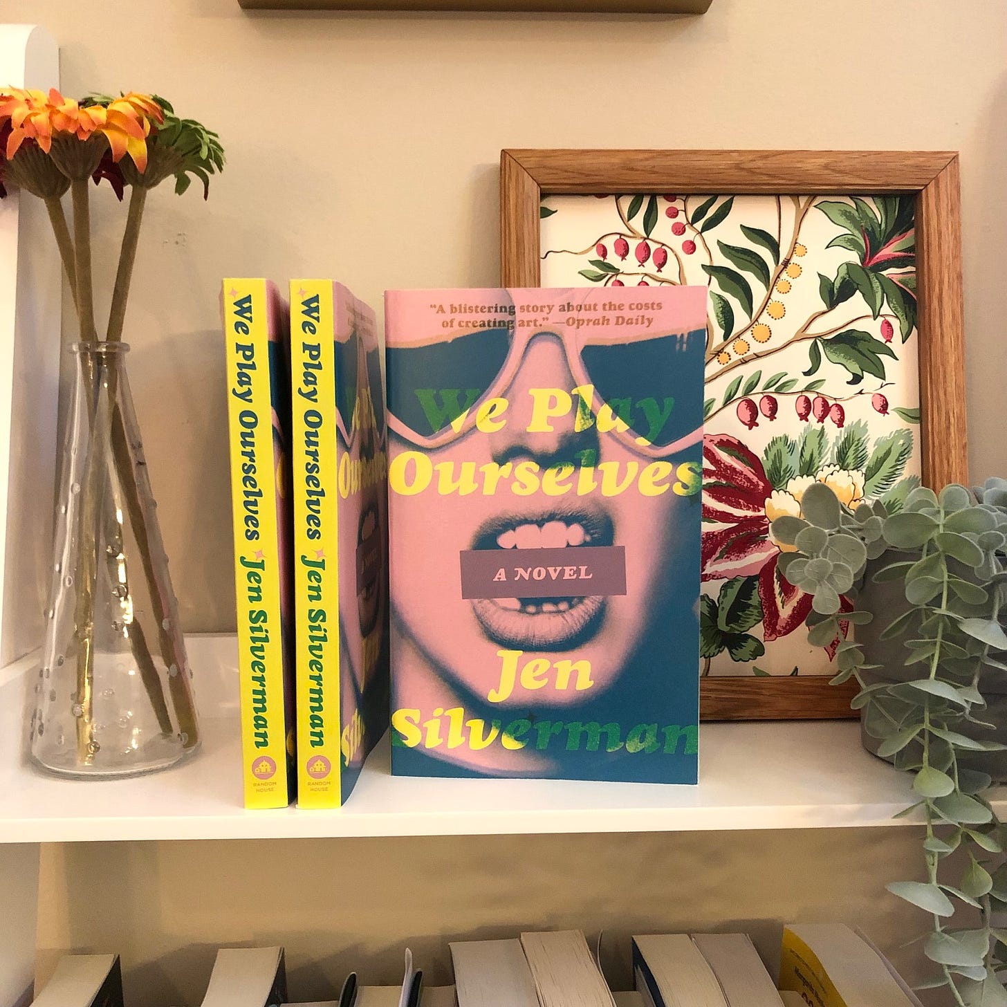 A close-up of three paperback copies of We Play Ourselves. The books are on a shelf with two fake plants and a framed floral painting behind them. 