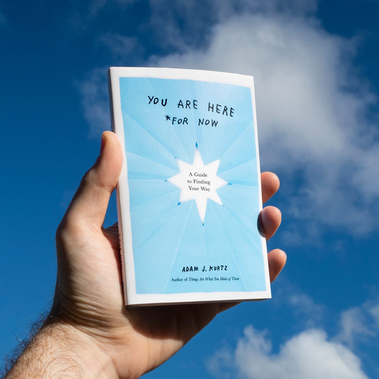 A white male hand holds the book You Are Here (For Now) against the sky—the cover looks like a blue paper cutout of a star with a white border
