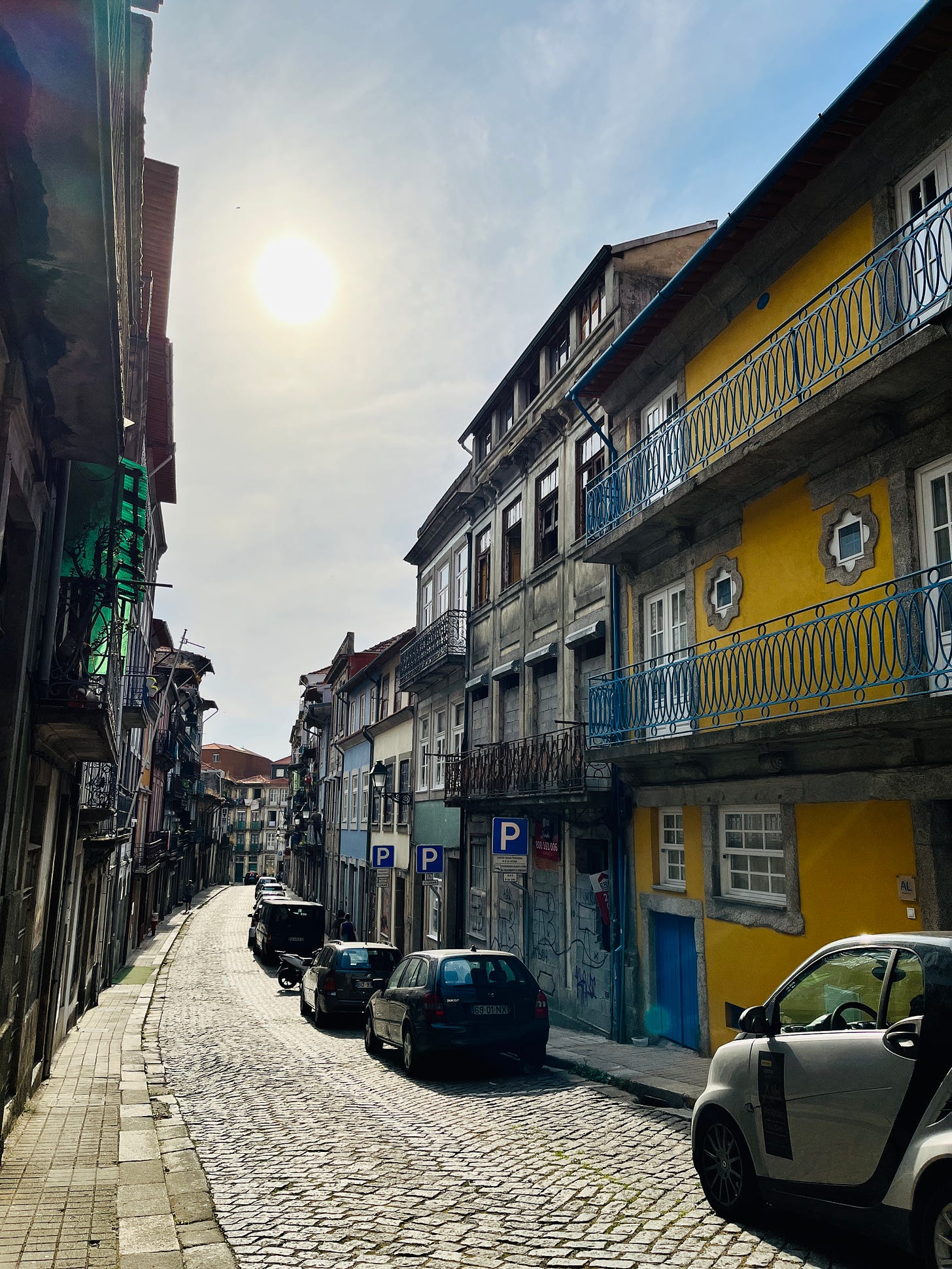 Image: a quiet inner street of Porto. Rows of very old Porto houses, opposite each other, in the middle, a cobblestone street curving downwards. The silhouette of the summer sun hangs proudly above. 