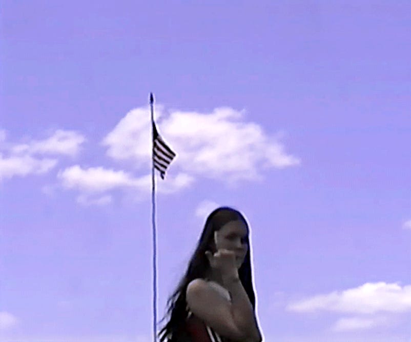 Ethel Cain, a woman with long brown hair and wearing a red and white cheerleading uniform, stands in front of an American flag raised on a flagpole with the cloudy blue sky behind it. She is giving the camera the middle finger.