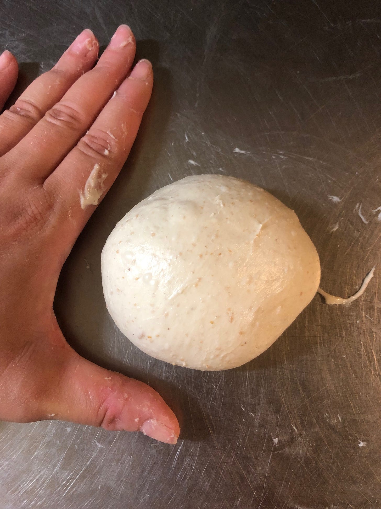 Left hand, palm down, on a metal table, next to a piece of raw dough shaped into a roll.