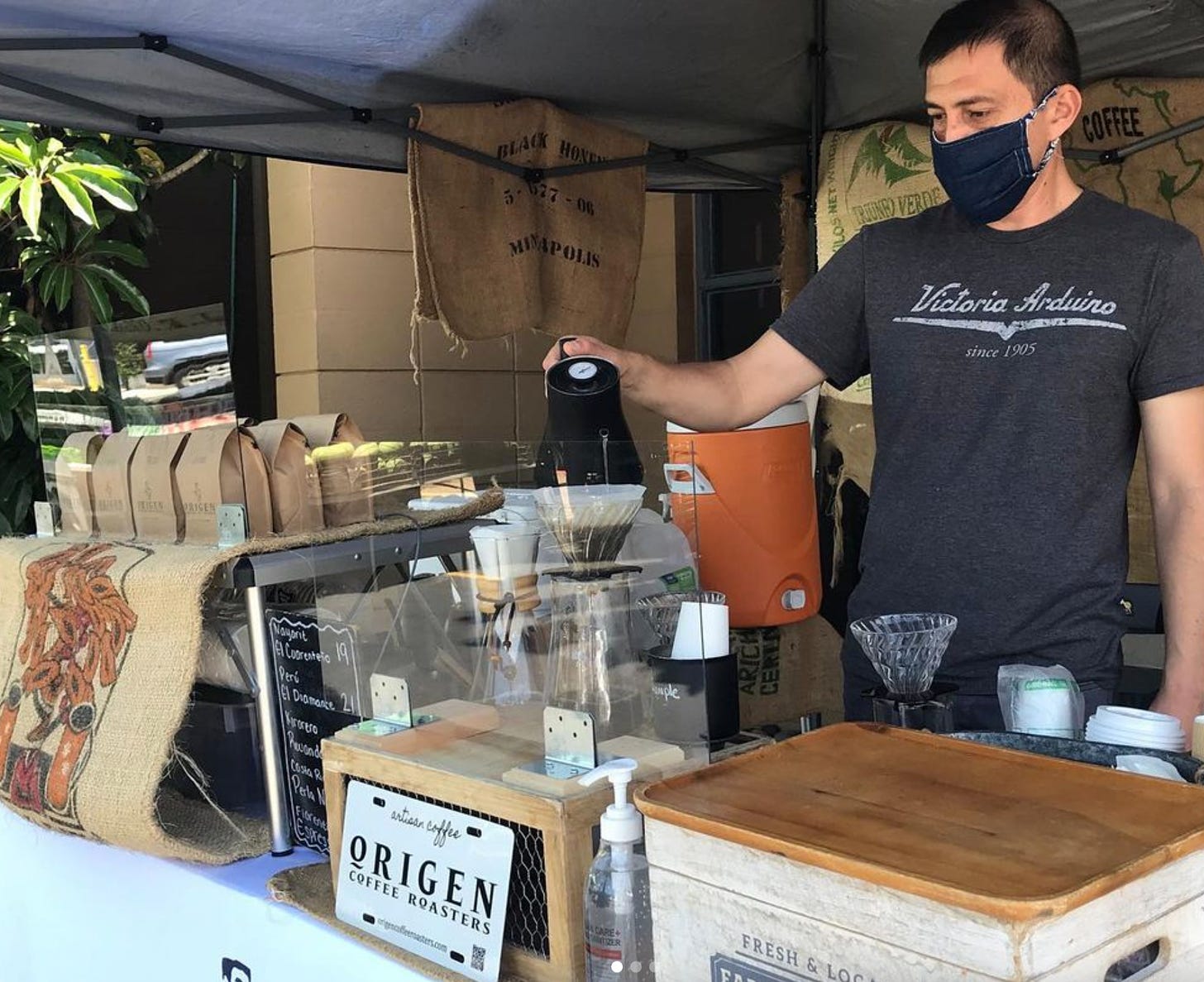 Ruben Enriquez pouring hot water into a conical pour over device holding coffee grounds under the pop-up tent and in the booth at a local farmers market. Coffee bags with beans for sale and coffee equipment line the table. 