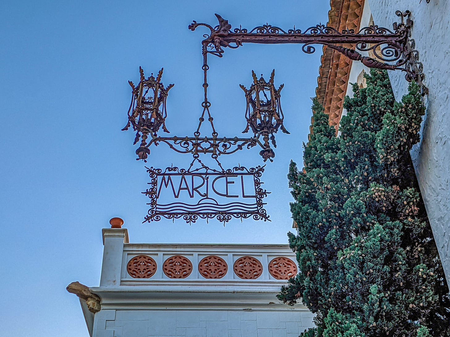 The elaborate wrought-iron sign for Museu de Maricel hanging from the side of a building and including two elaborate lamps. 
